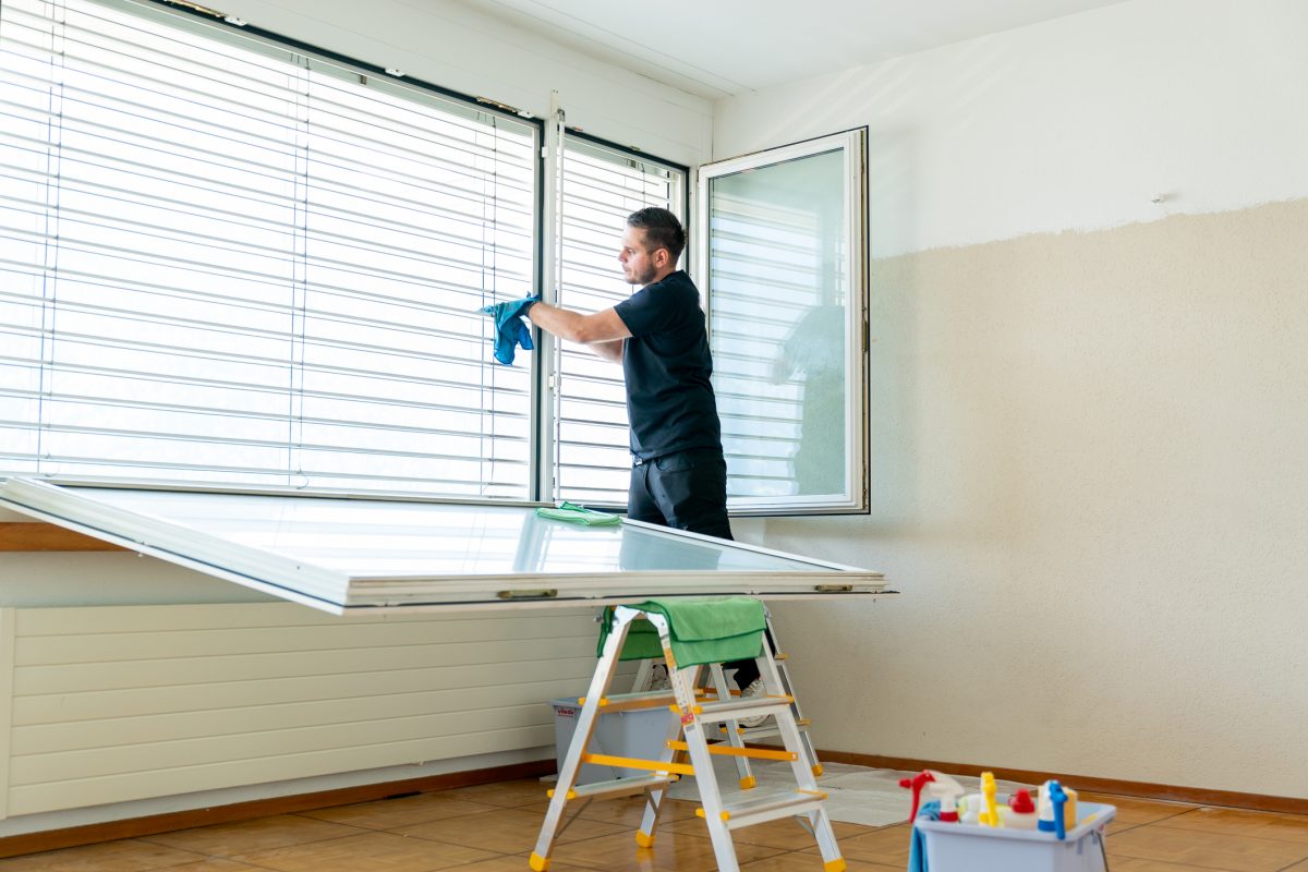 A professional cleaner cleaning window blinds in an apartment with a blue micro fiber cloth